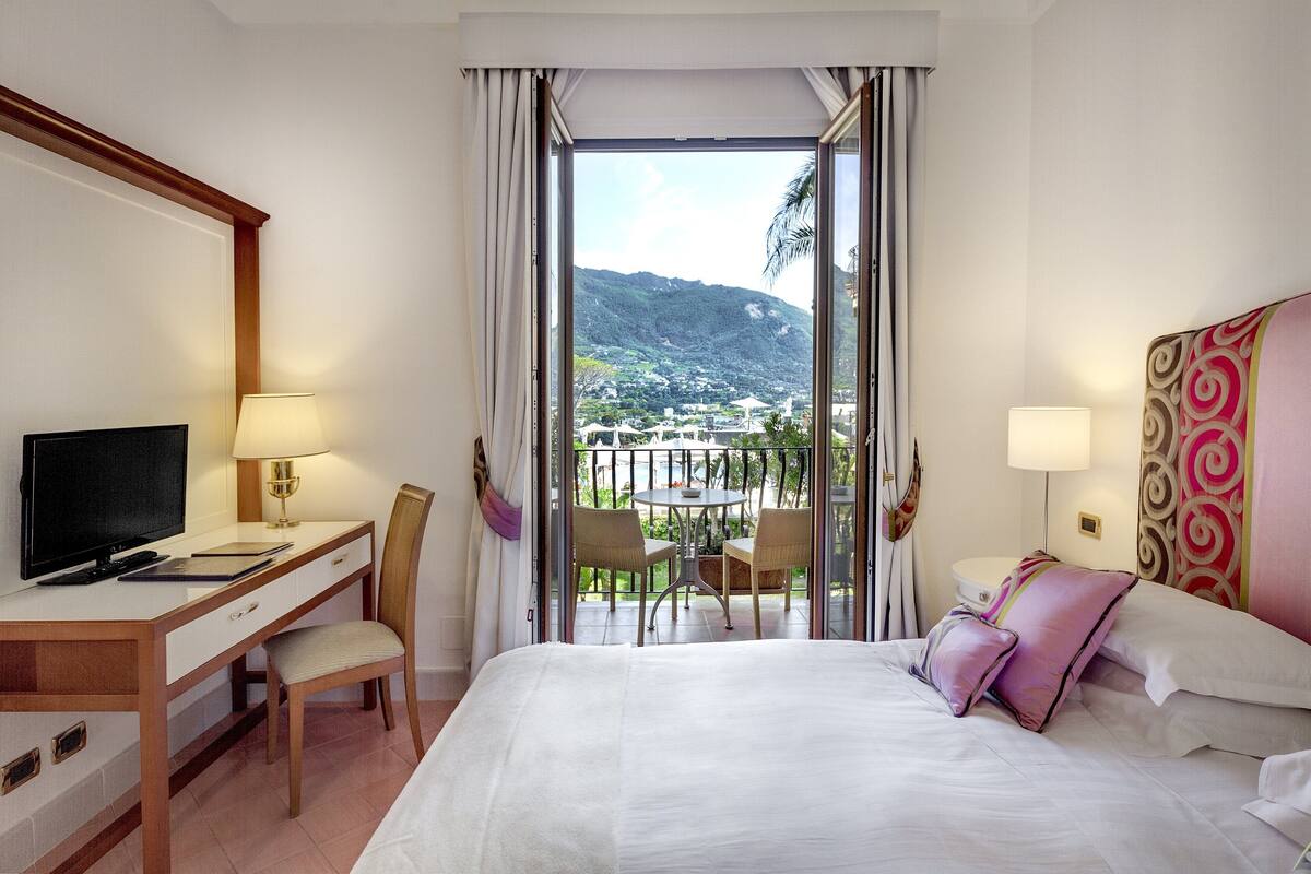 Ein Hotelzimmer mit Bett, Schreibtisch mit Computer, Fernseher und Balkon mit Stühlen und Tisch.