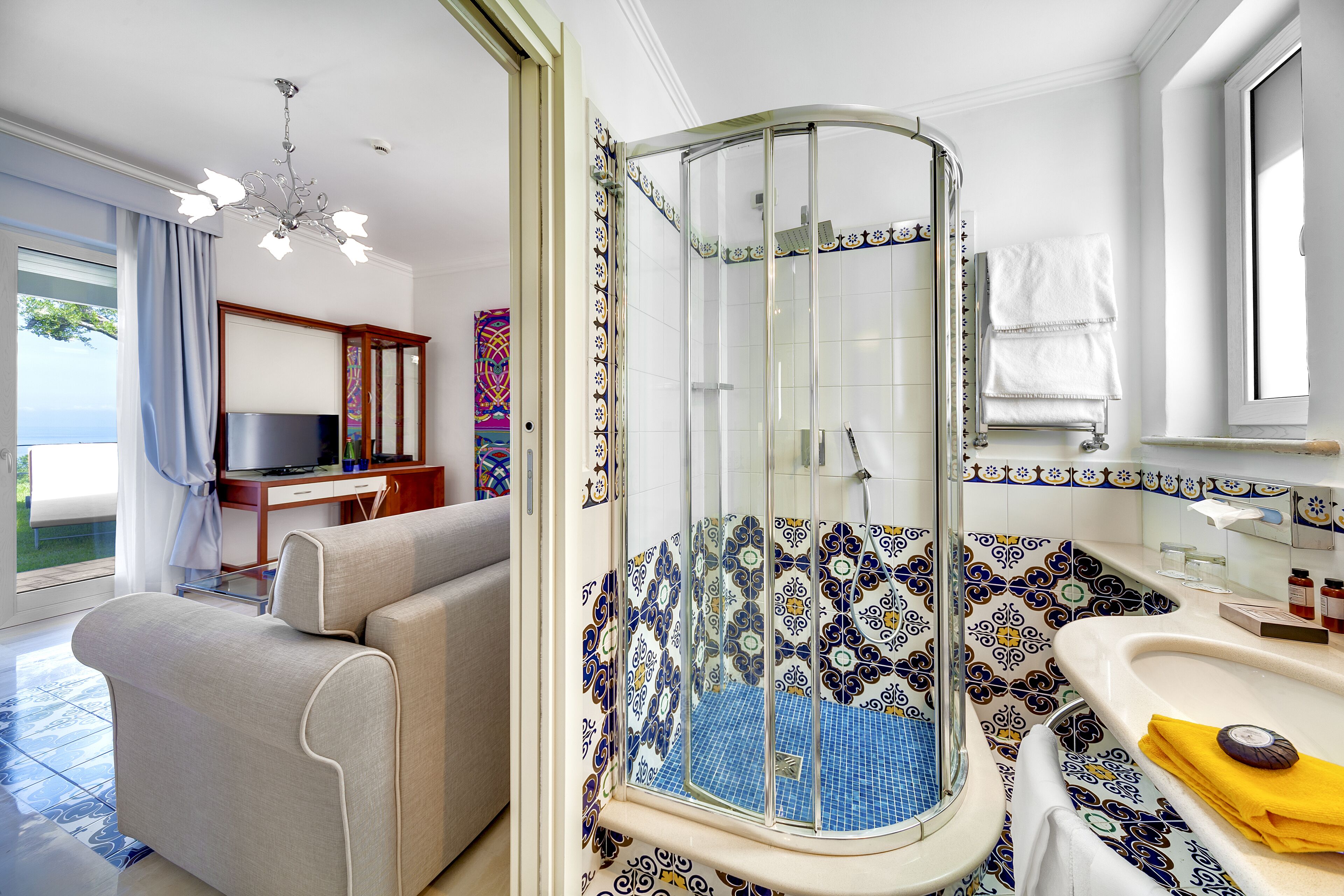 A modern bathroom with a glass-enclosed shower, a beige sofa, and a television in the living area.