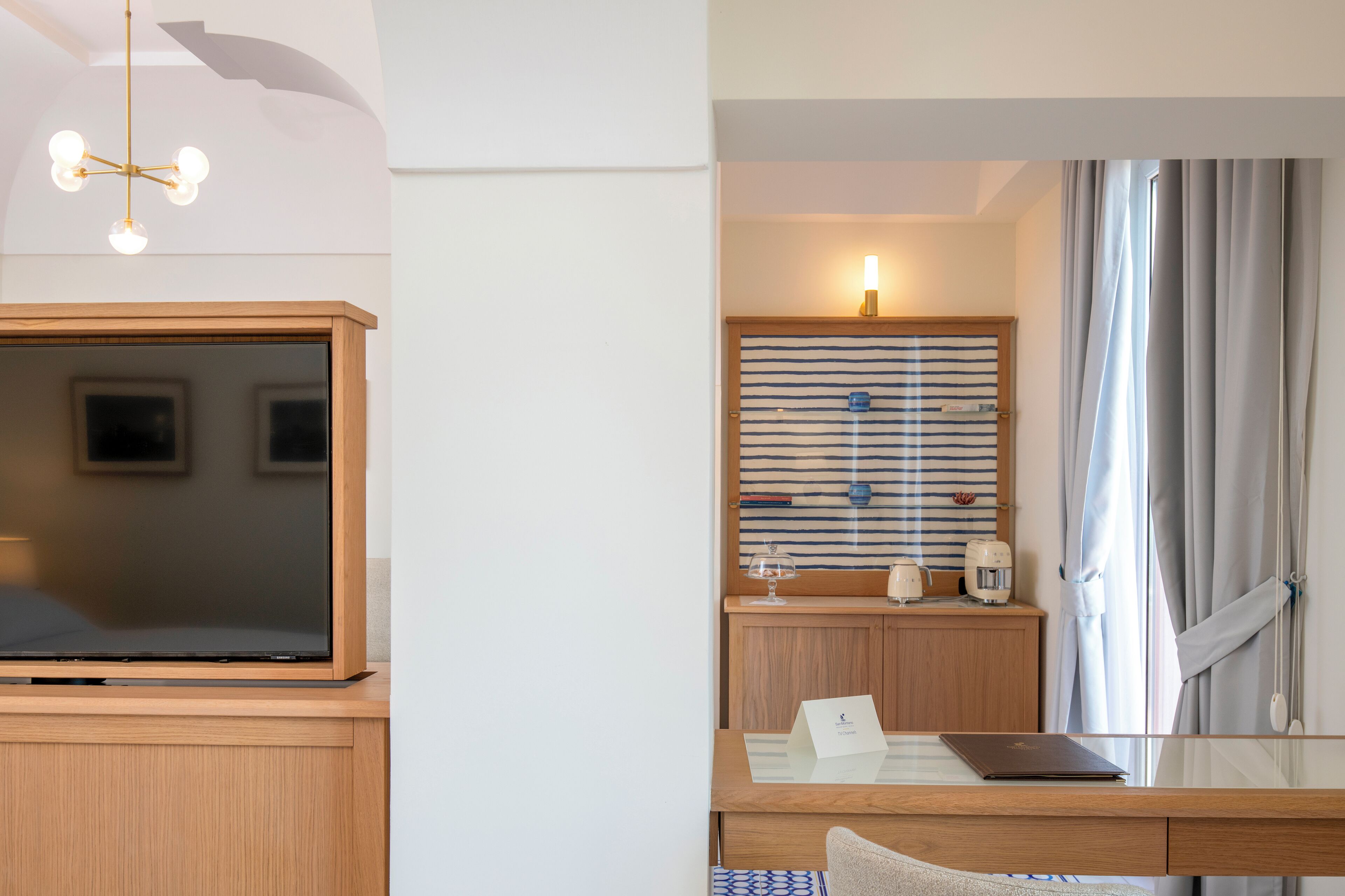 A hotel room with a wooden desk, a television, a striped wall, and a window with curtains.