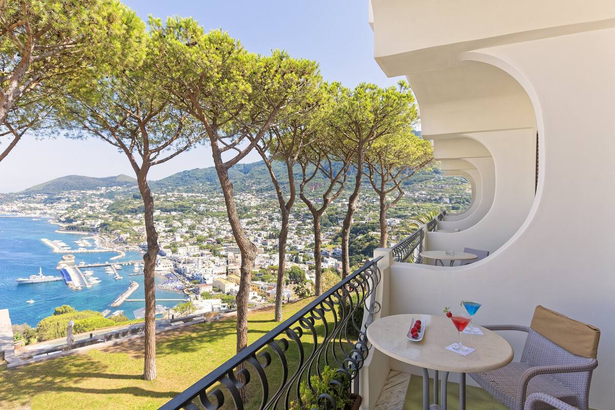 Ein Balkon mit Blick auf das Meer und die Stadt, mit einem Tisch, einem Getränk und einer Fruchtschale.