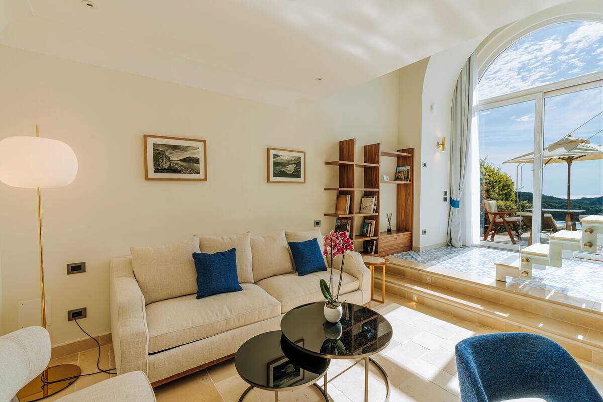 Ein modernes Wohnzimmer mit einer Couch, einem Couchtisch und einem Bücherregal. Ein großes Fenster bietet einen Blick ins Freie.