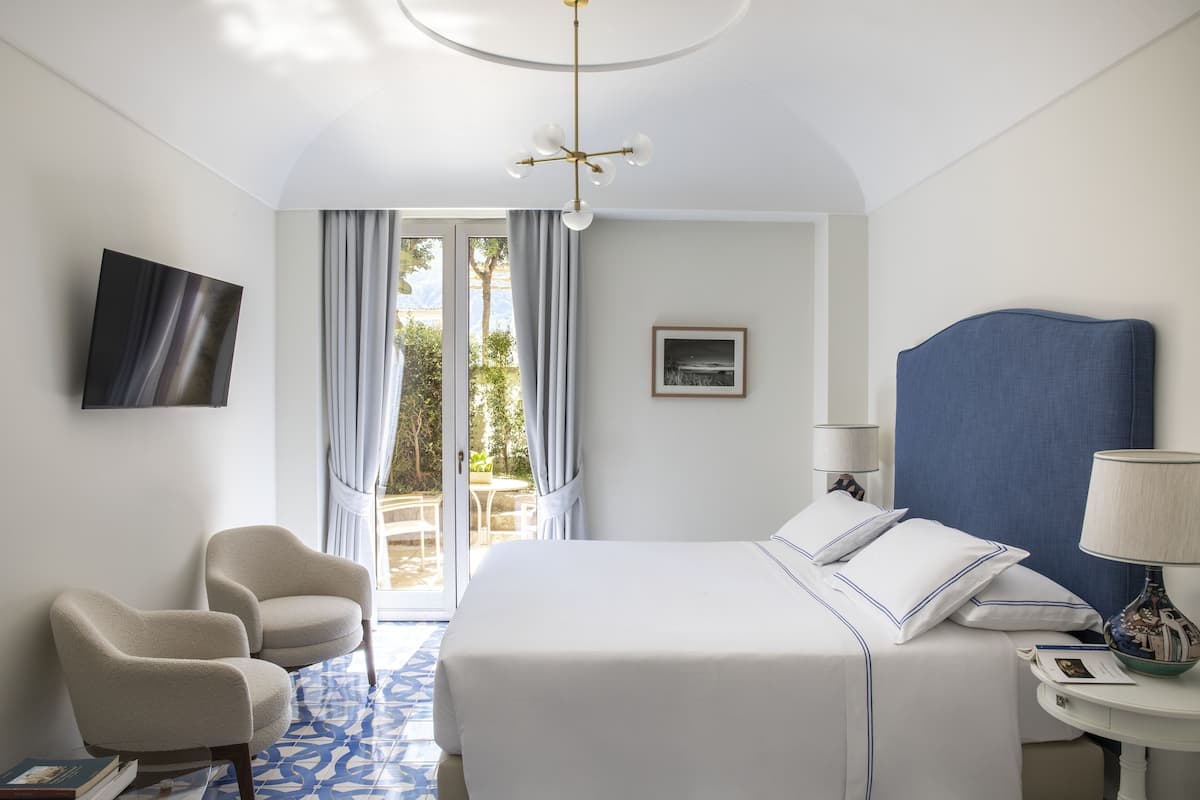 Ein Hotelzimmer mit einem Bett, zwei Sesseln, einem Fernseher, einer Lampe und Blick ins Freie.