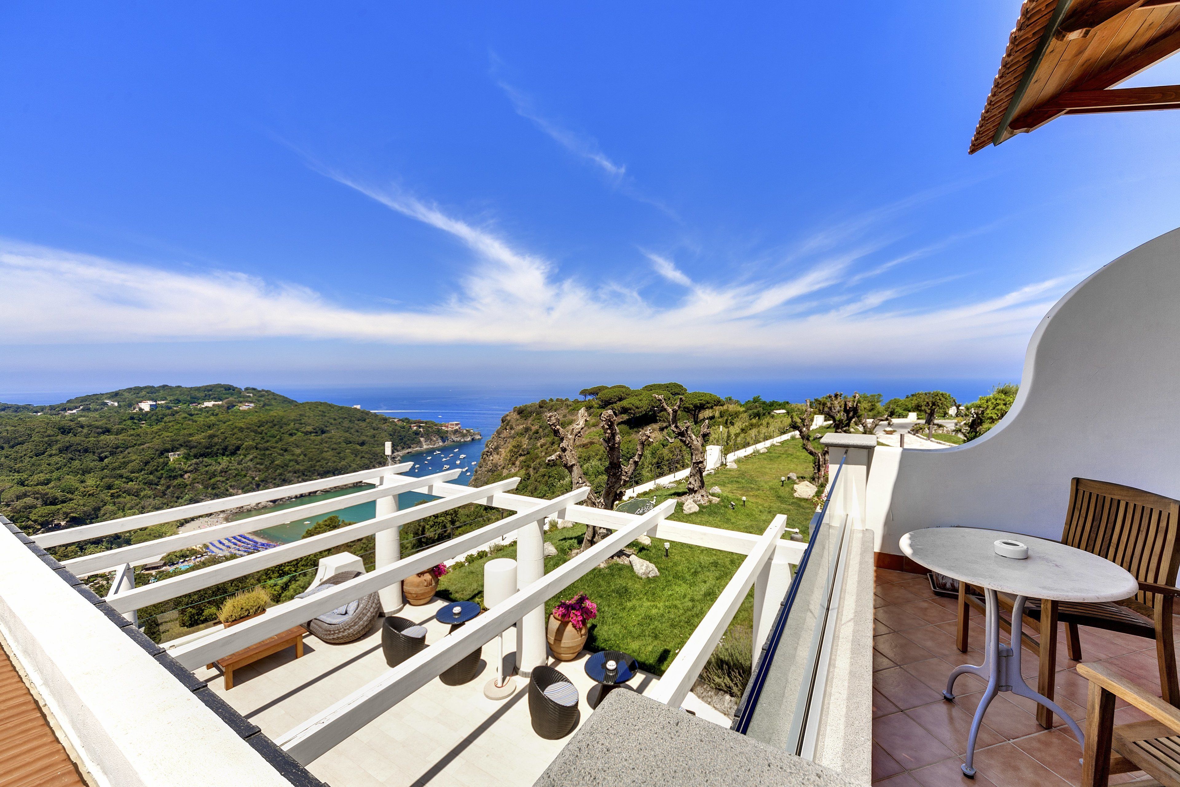 A terrace with a view of the sea and hills, furnished with a table and chairs, and a white railing.