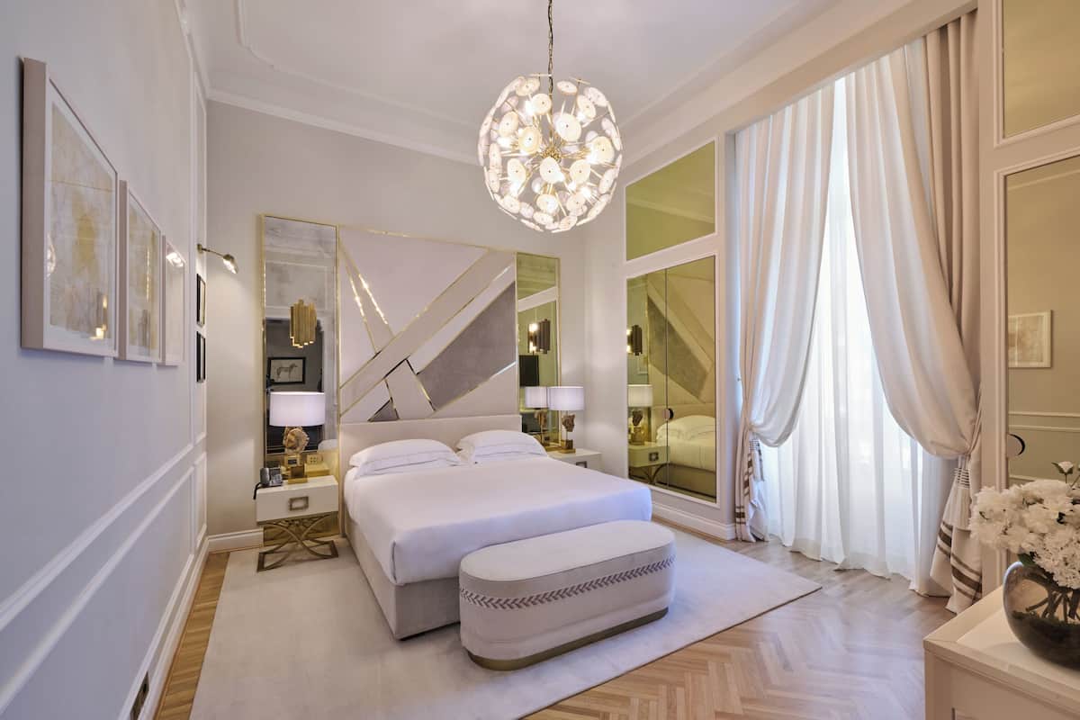A modern bedroom with a large bed, a mirror, a chandelier, and a view of the city through floor-to-ceiling windows.
