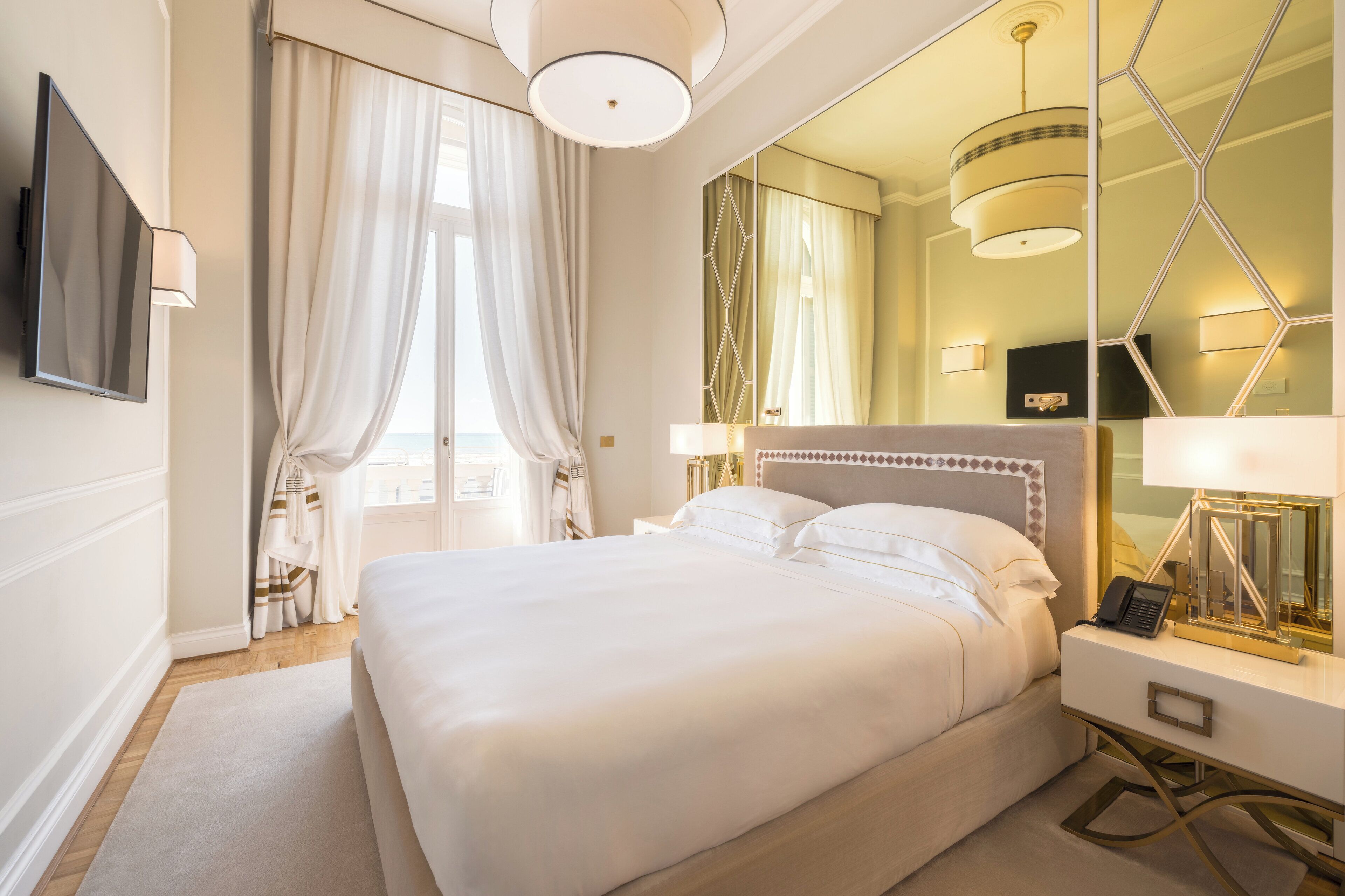 Hotelkamer met een groot bed, nachtkastje met een lamp, flatscreen televisie aan de muur en een grote spiegel met een geometrisch frame.