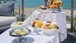 A table set with breakfast items, including oysters, croissants, and a bottle of champagne, overlooking a beach.
