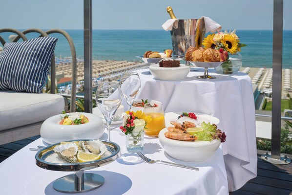 A table set with breakfast items, including oysters, croissants, and a bottle of champagne, overlooking a beach.