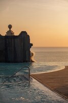 A wooden deck leading to a pool with a clear view of the ocean and a rocky cliff.