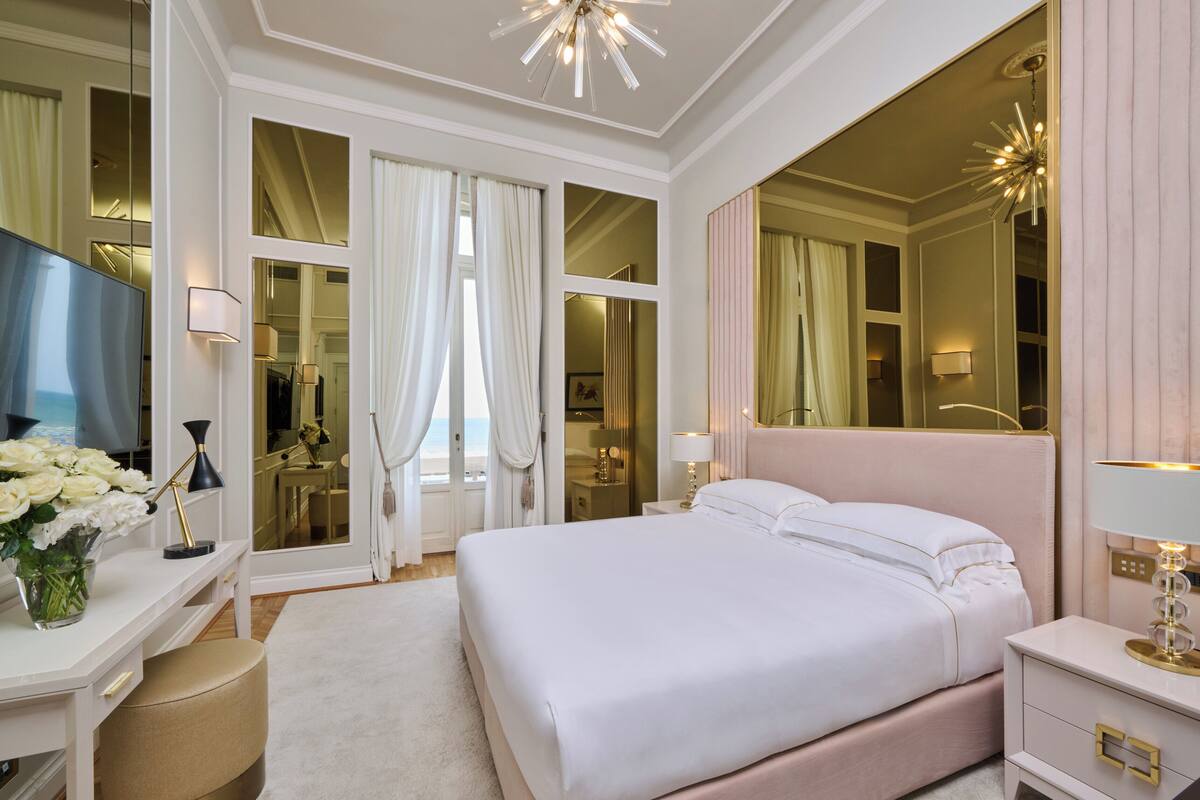 A spacious bedroom with a large bed, a desk with a lamp, a vanity with a mirror, and a large window with curtains.