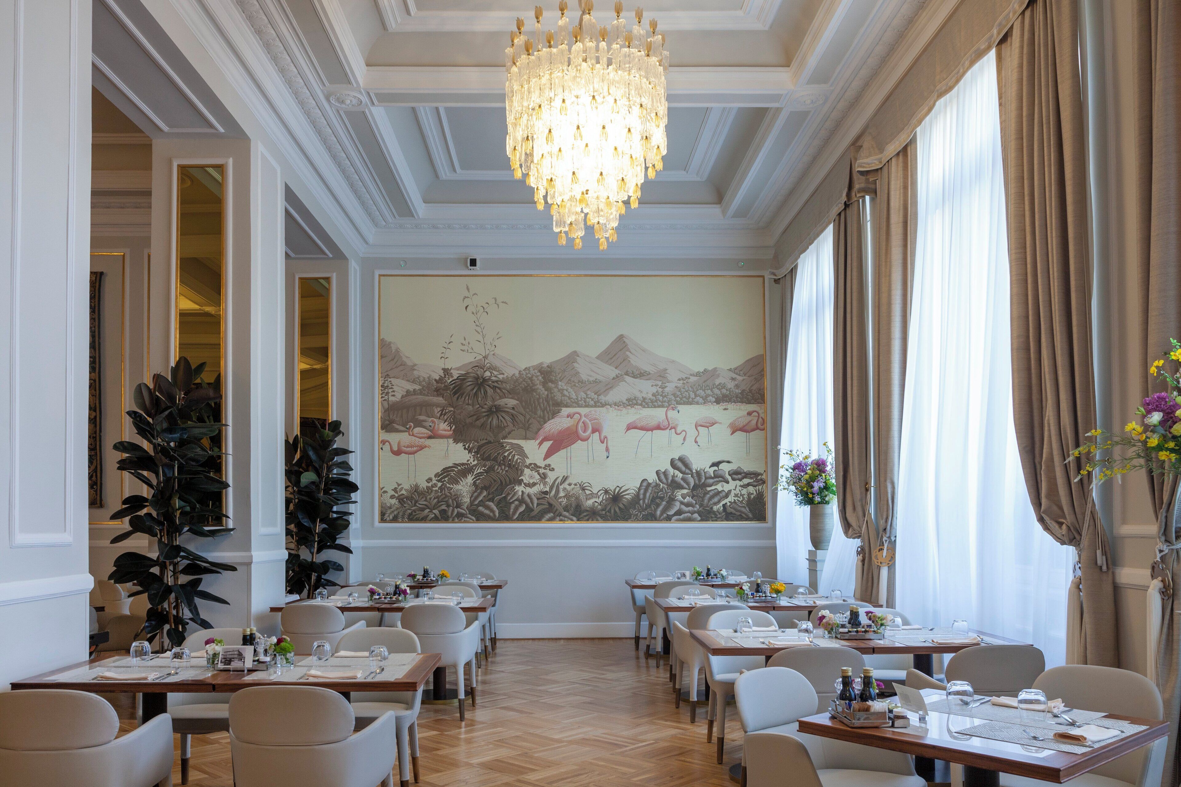 Un comedor con mesas dispuestas para el servicio, una araña de cristal y un mural grande en la pared.