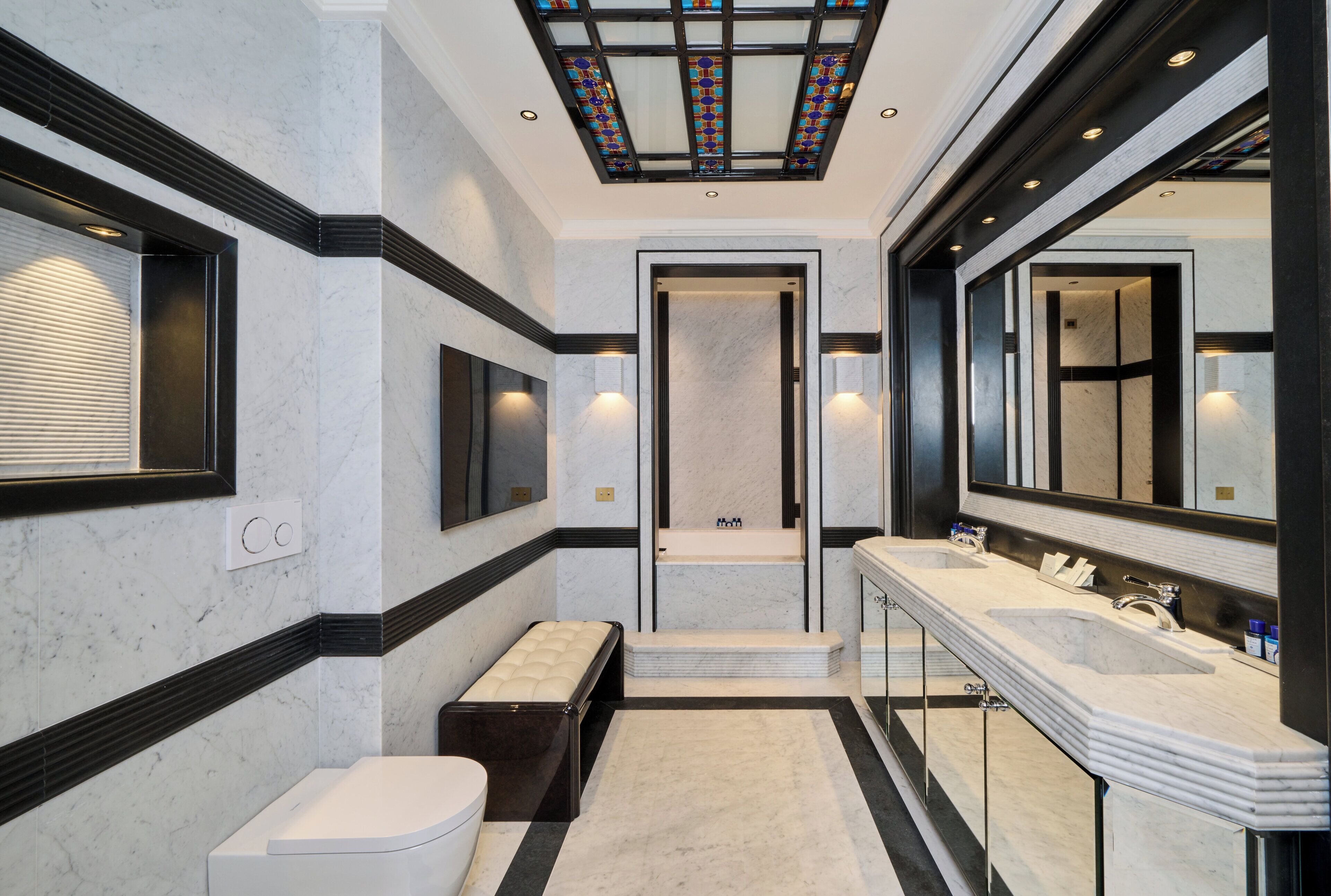 A modern bathroom with a large mirror, two sinks, a bench, and a shower area.