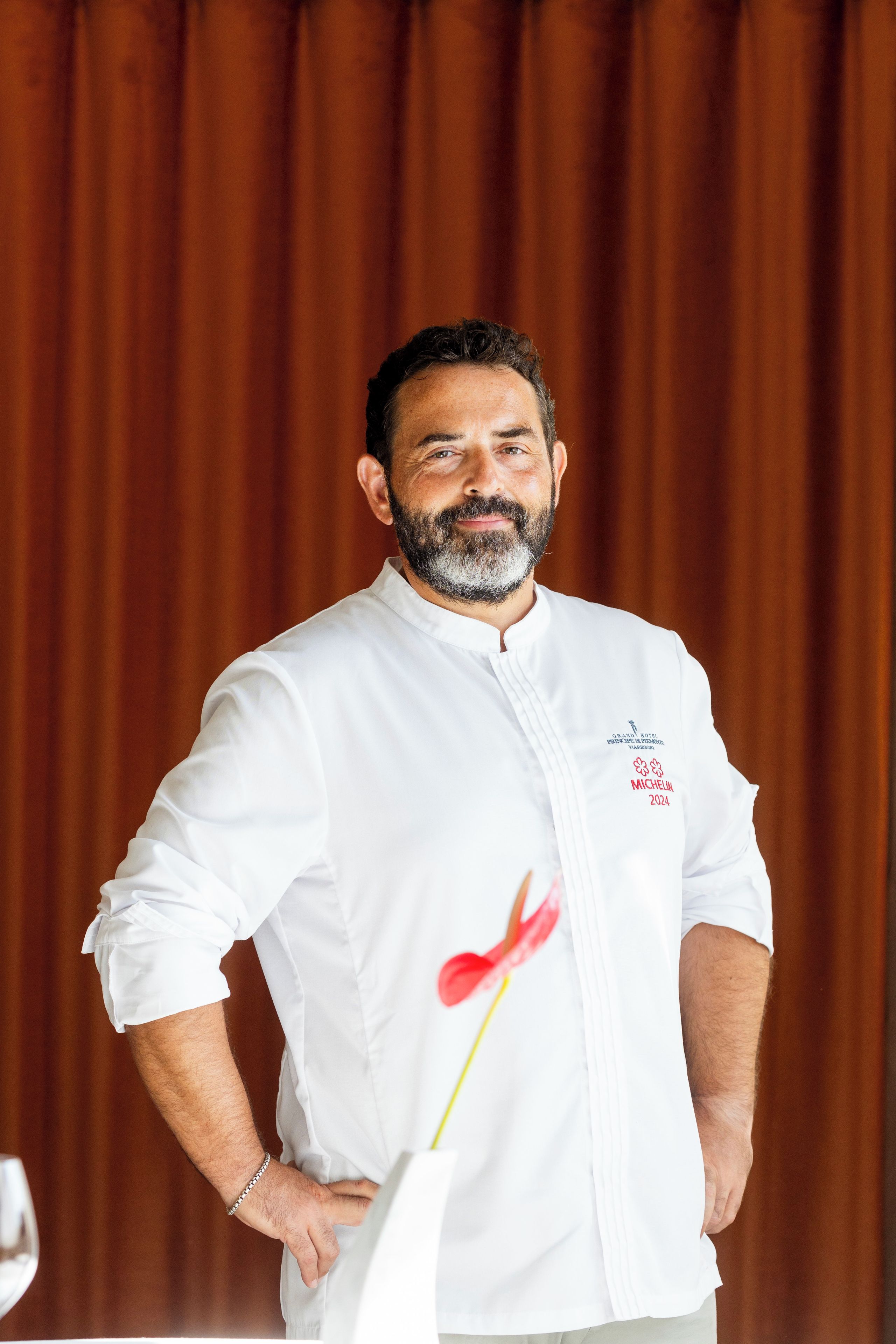 Una persona con bata de chef blanca con un logo y texto en el lado izquierdo, de pie frente a una cortina marrón.