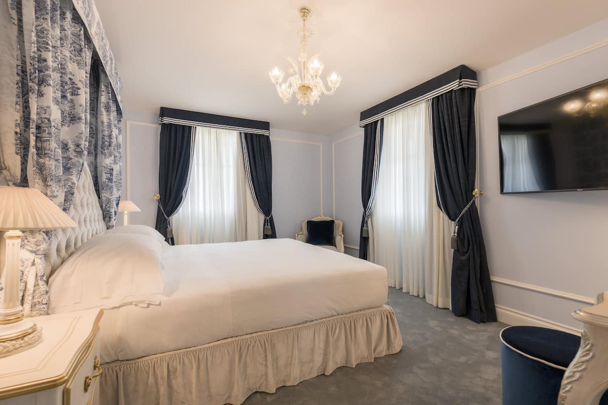 A hotel room with a large bed, a chandelier, a flat-screen TV, and a classic chair.