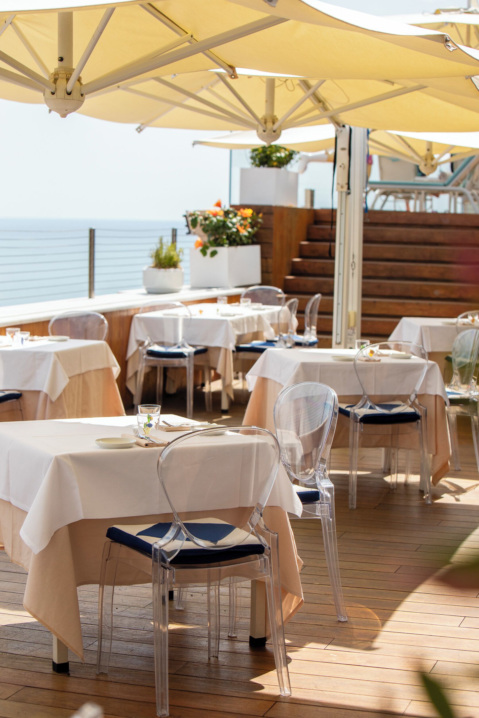 Tavoli da pranzo all'aperto con sedie trasparenti, tovaglie bianche e ombrelloni, con vista su un pontile di legno e sull'oceano.