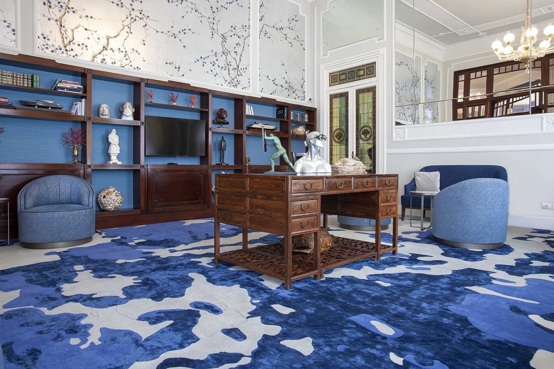 A room with a large wooden desk, blue armchairs, a patterned rug, and a bookshelf filled with books and decorative items.