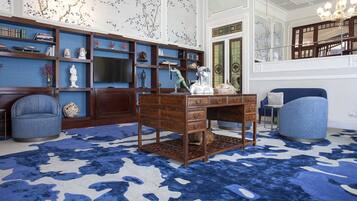 A room with a large wooden desk, blue armchairs, a patterned rug, and a bookshelf filled with books and decorative items.