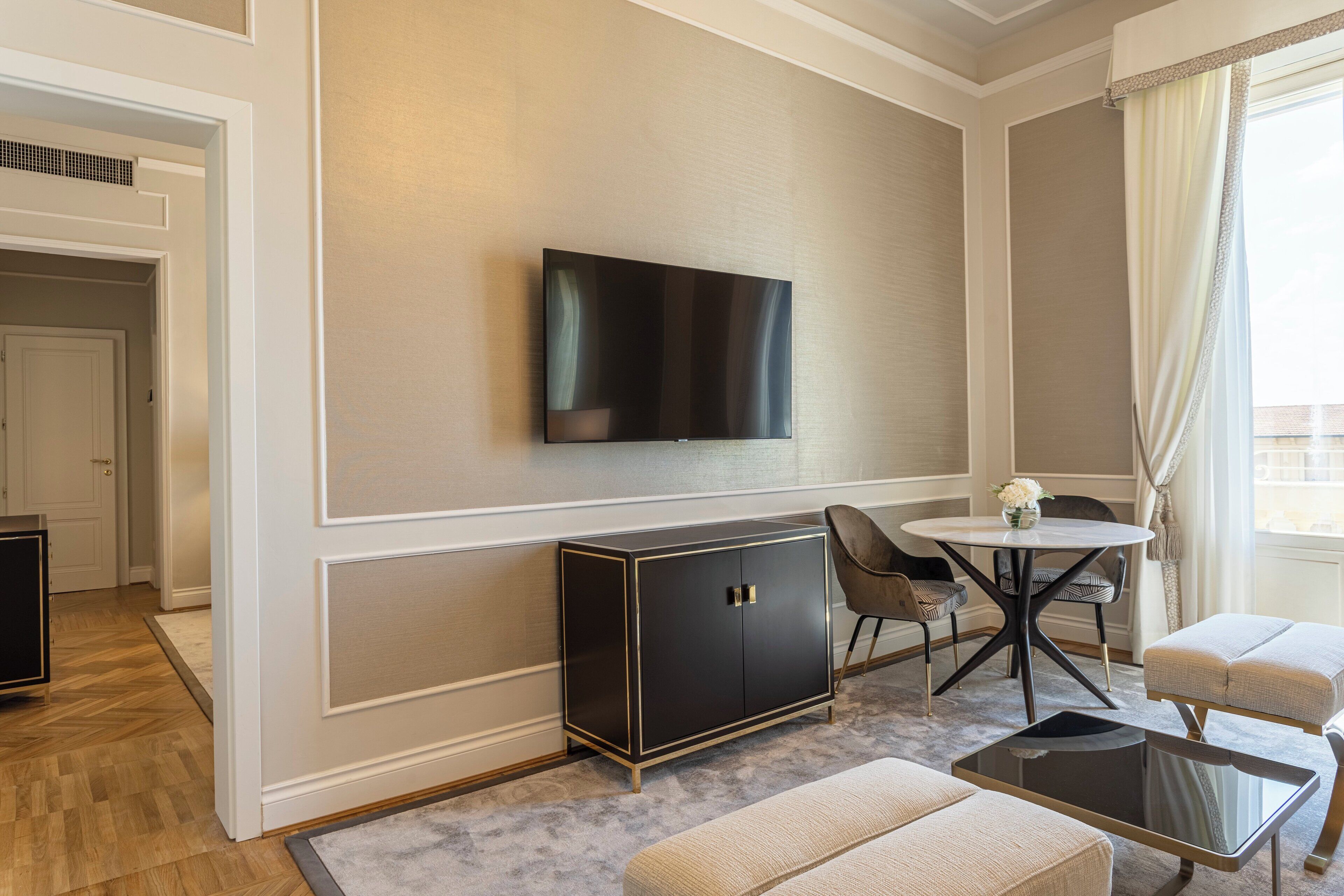 Un salon moderne avec un téléviseur à écran plat fixé au mur, un meuble en bois foncé, une table de salle à manger ronde avec des chaises et un petit pouf.