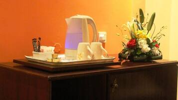 A hotel room with a coffee maker, two white cups, a tray with a small bowl and a spoon, and a floral arrangement on a wooden cabinet.