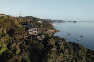 A coastal house with a large balcony overlooking the sea, surrounded by dense forest.