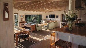 A spacious living area with a wooden ceiling, a dining table with chairs, a sofa, and a kitchen area with a counter and a vase of flowers. - Cliff Edge by the Sea (Opua)