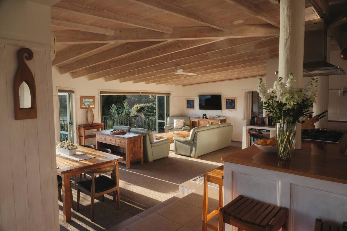A spacious living area with a wooden ceiling, a dining table with chairs, a sofa, and a kitchen area with a counter and a vase of flowers.