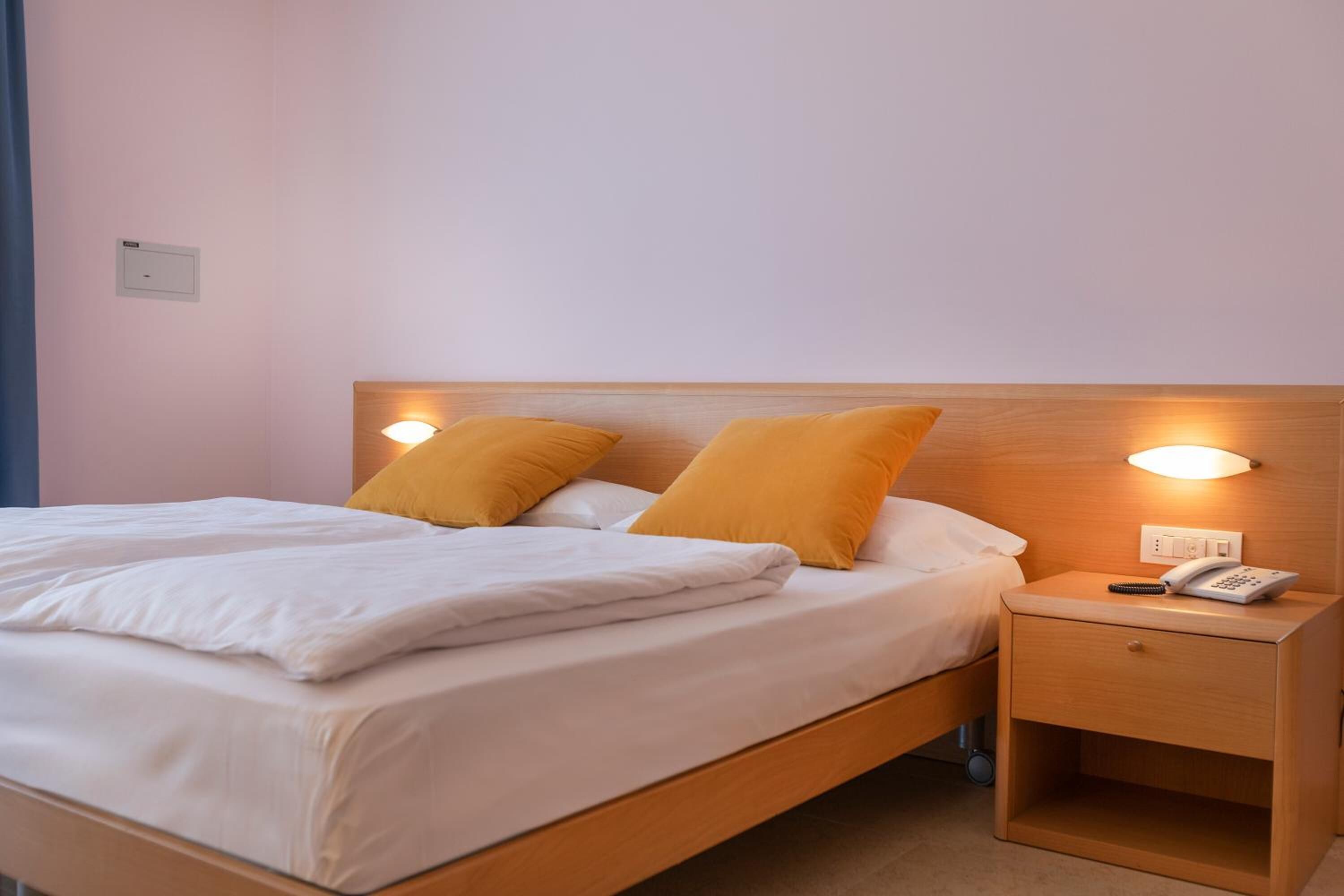 Un lit double avec une literie blanche et deux oreillers orange, une table de chevet en bois avec un téléphone, et une lampe fixée au mur.