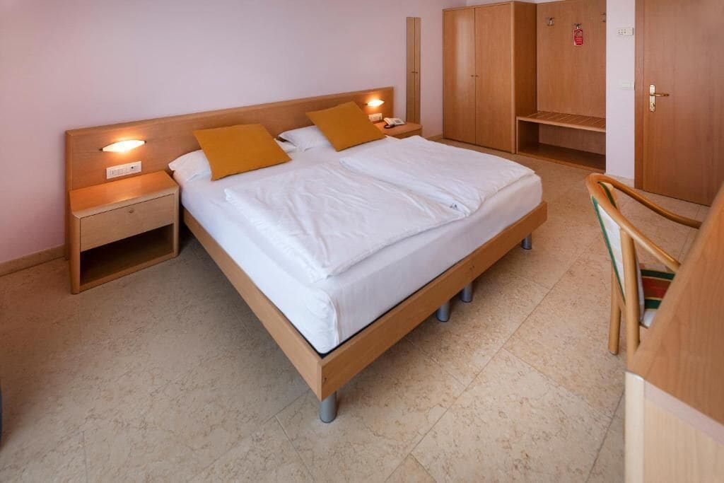 Habitación individual con una cama de madera, dos mesitas de noche, un armario y una silla.