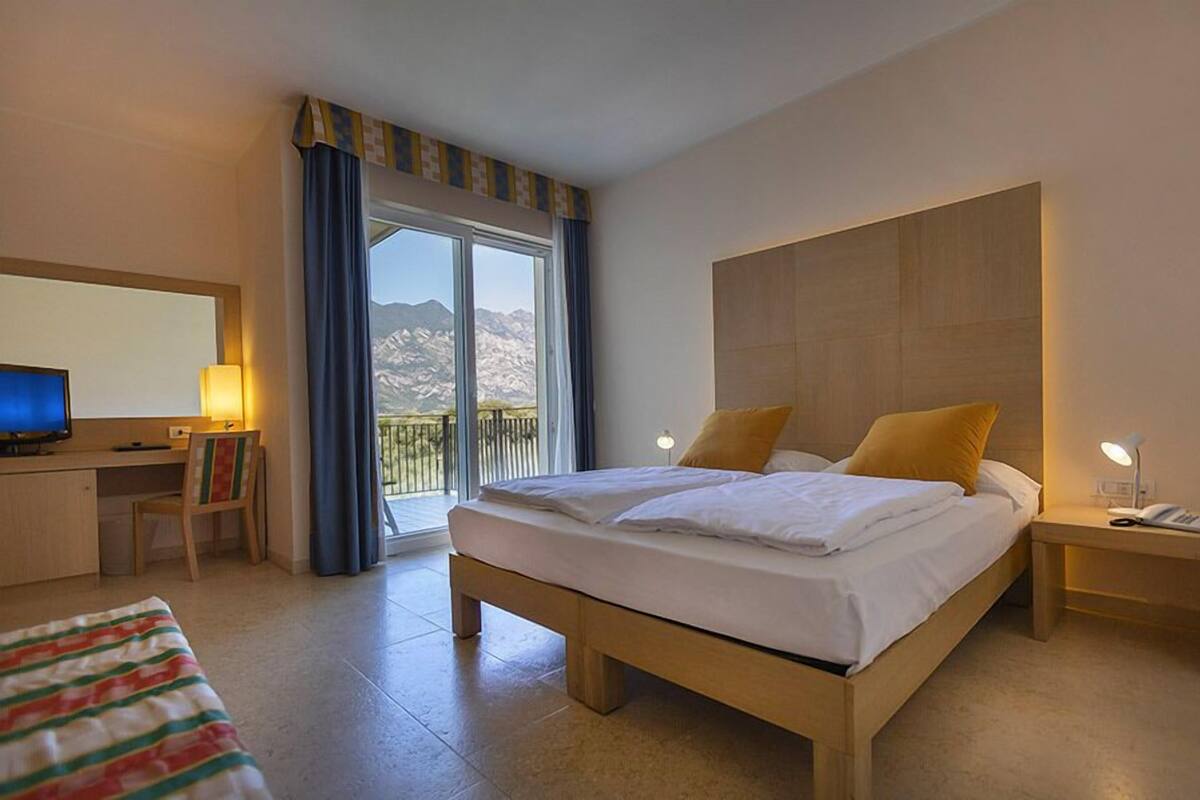 Une chambre d’hôtel avec un lit, un bureau équipé d’un ordinateur, une chaise, une table de chevet avec une lampe, et une vue sur les montagnes par une grande fenêtre.