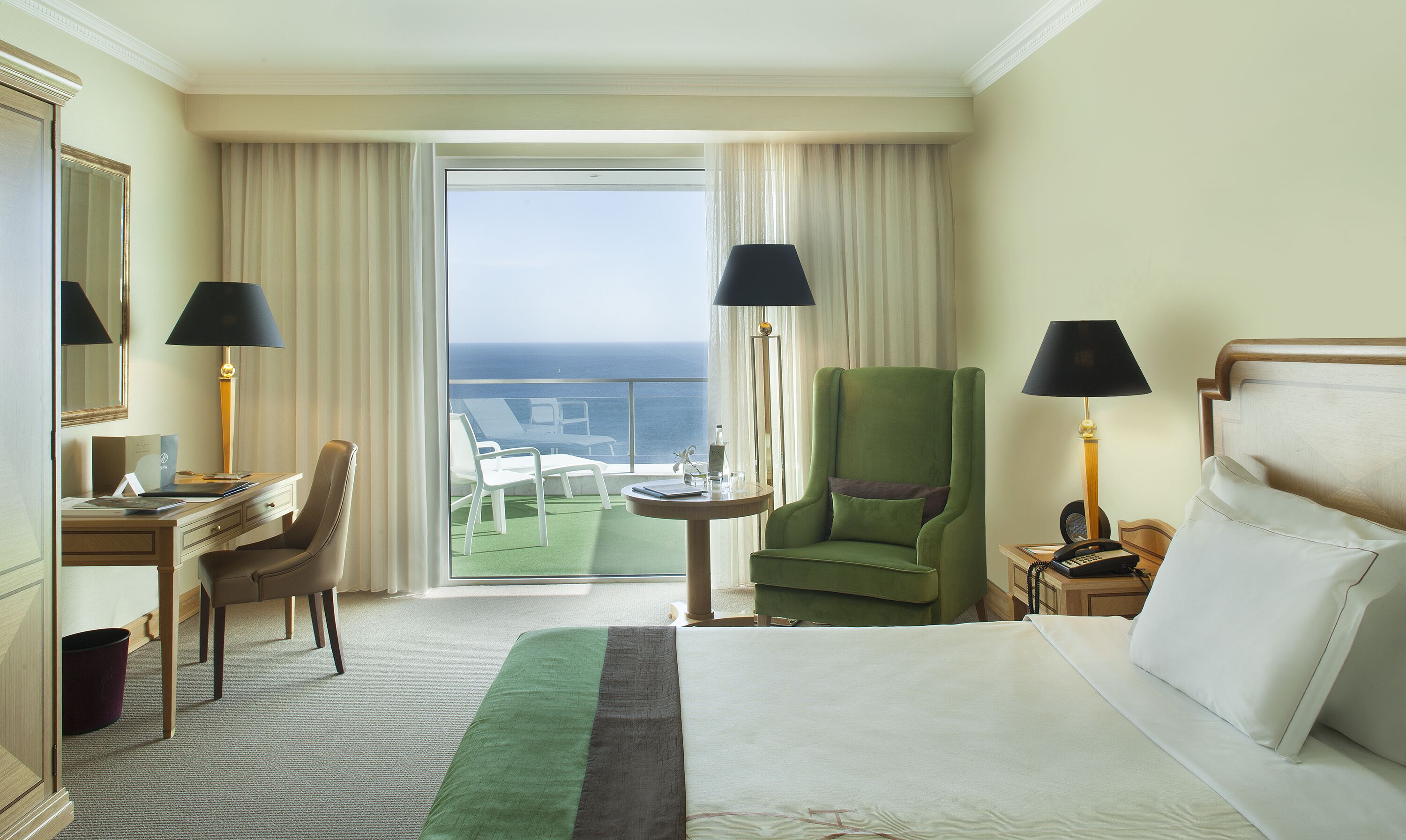 Un cuarto de hotel con una cama, un escritorio, una silla, un sillón verde y vista al océano.