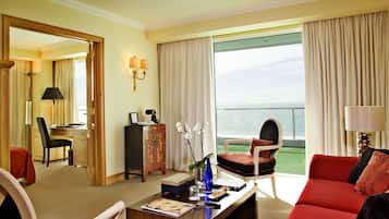 A hotel room with a large window, a red sofa, a dining area with a table and chairs, and a desk with a computer and lamp.