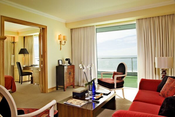 A hotel room with a large window, a red sofa, a dining area with a table and chairs, and a desk with a computer and lamp.