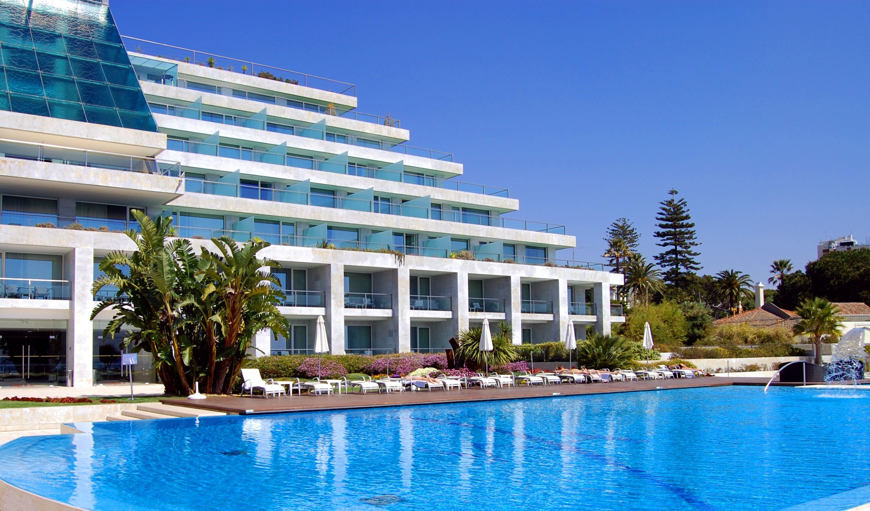 A modern hotel with a large swimming pool, multiple balconies, and lush landscaping.