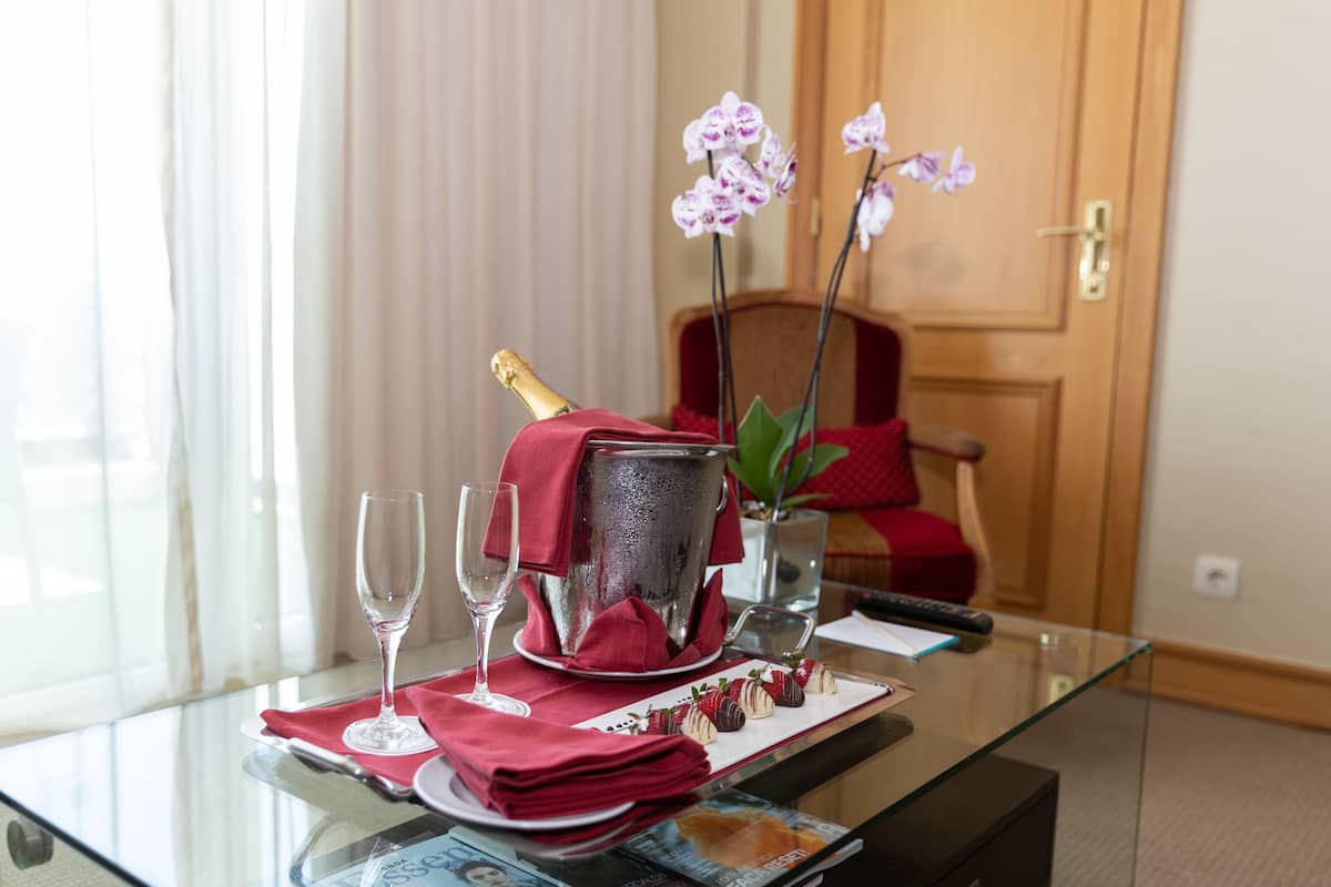 Una mesa de vidrio con una botella de champán, dos copas de vino, un paño de color rojo, un plato con fresas cubiertas de chocolate y una jarra con orquídeas.