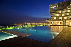 An infinity pool with a wooden deck, a modern building with balconies, and a cityscape view at dusk.