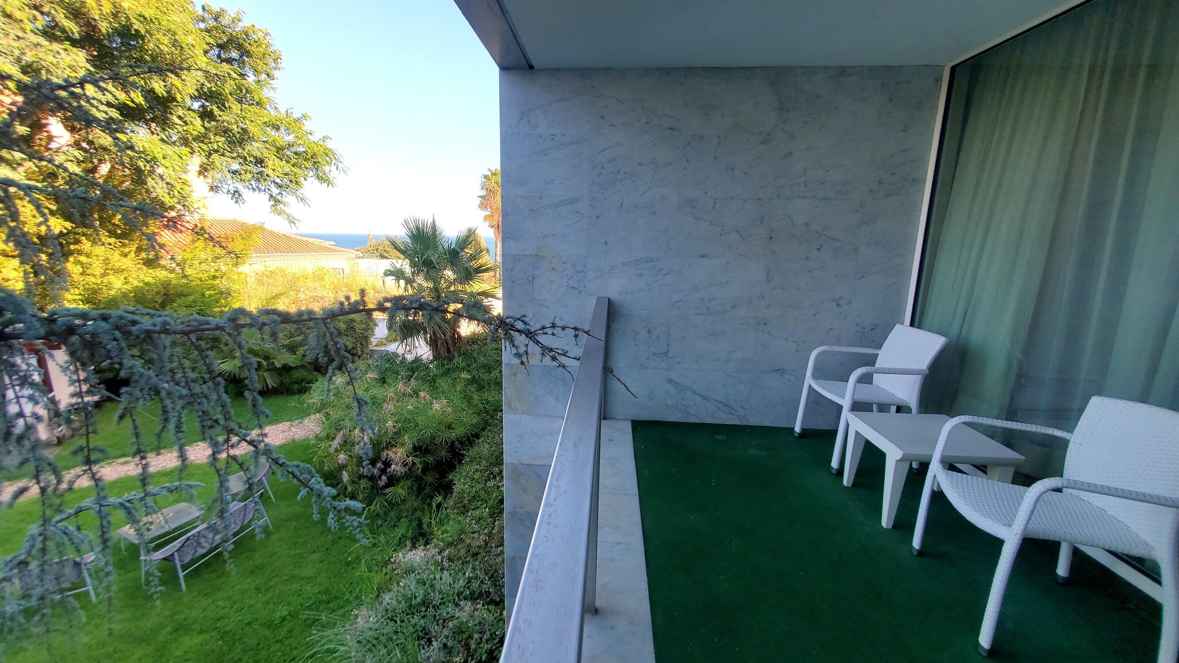 Un balcon agrémenté d’un gazon synthétique vert, de meubles en osier blancs et offrant une vue sur un jardin avec des arbres et un bâtiment en arrière-plan.