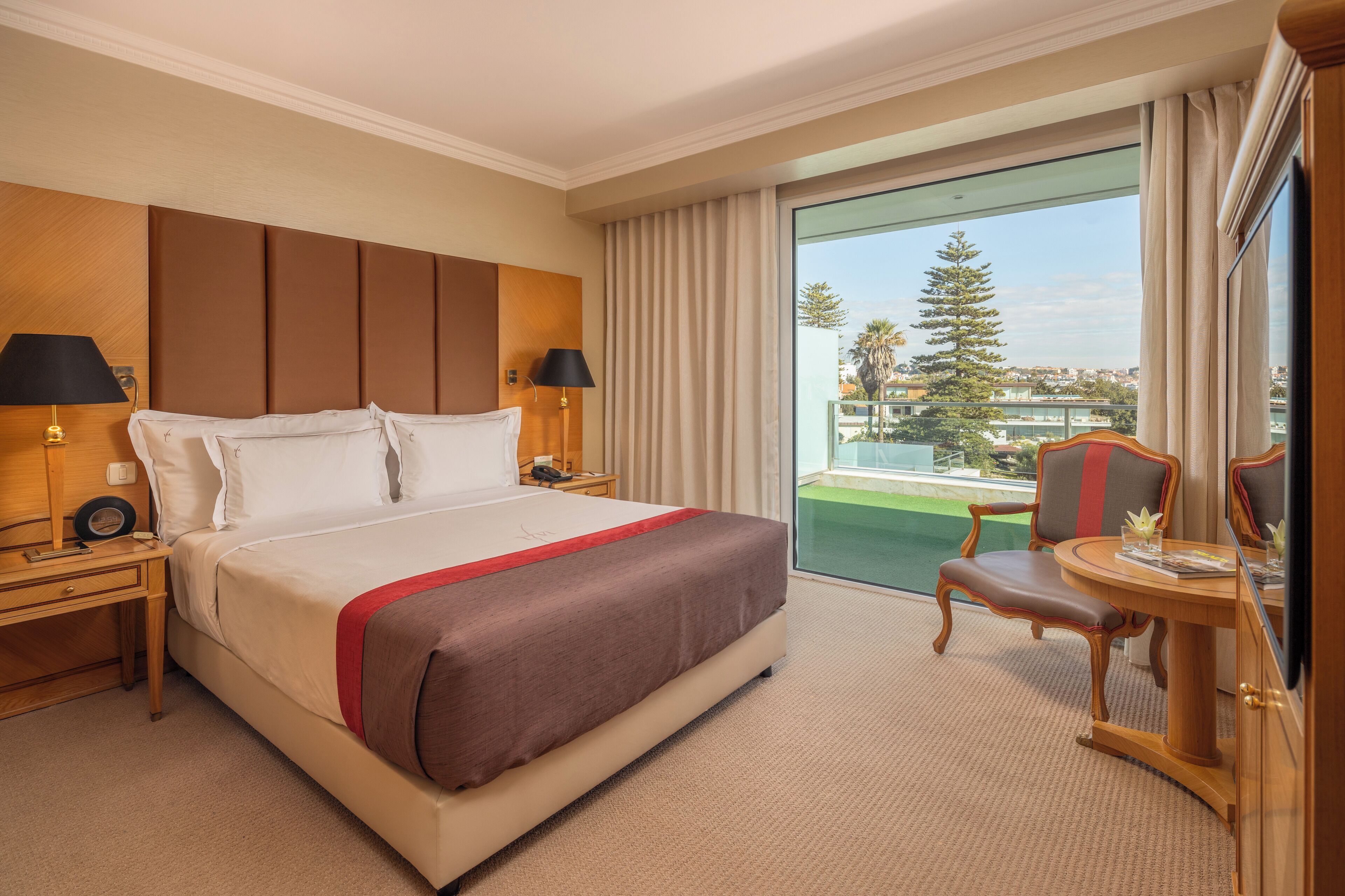 A hotel room with a large bed, two bedside tables with lamps, a small table with a chair, and a balcony with a view of the city and a tree.