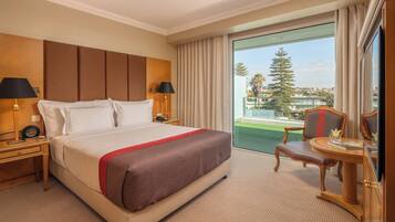 A hotel room with a large bed, two bedside tables with lamps, a small table with a chair, and a balcony with a view of the city and a tree.