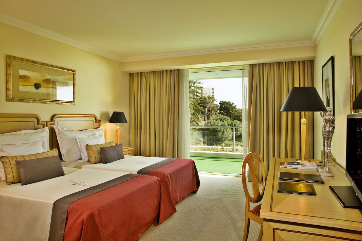 Habitación de hotel con dos camas, un escritorio con lámpara y vista a zonas verdes por la ventana.