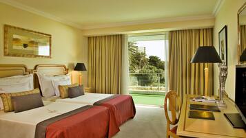Habitación de hotel con dos camas, un escritorio con lámpara y vista a zonas verdes por la ventana.