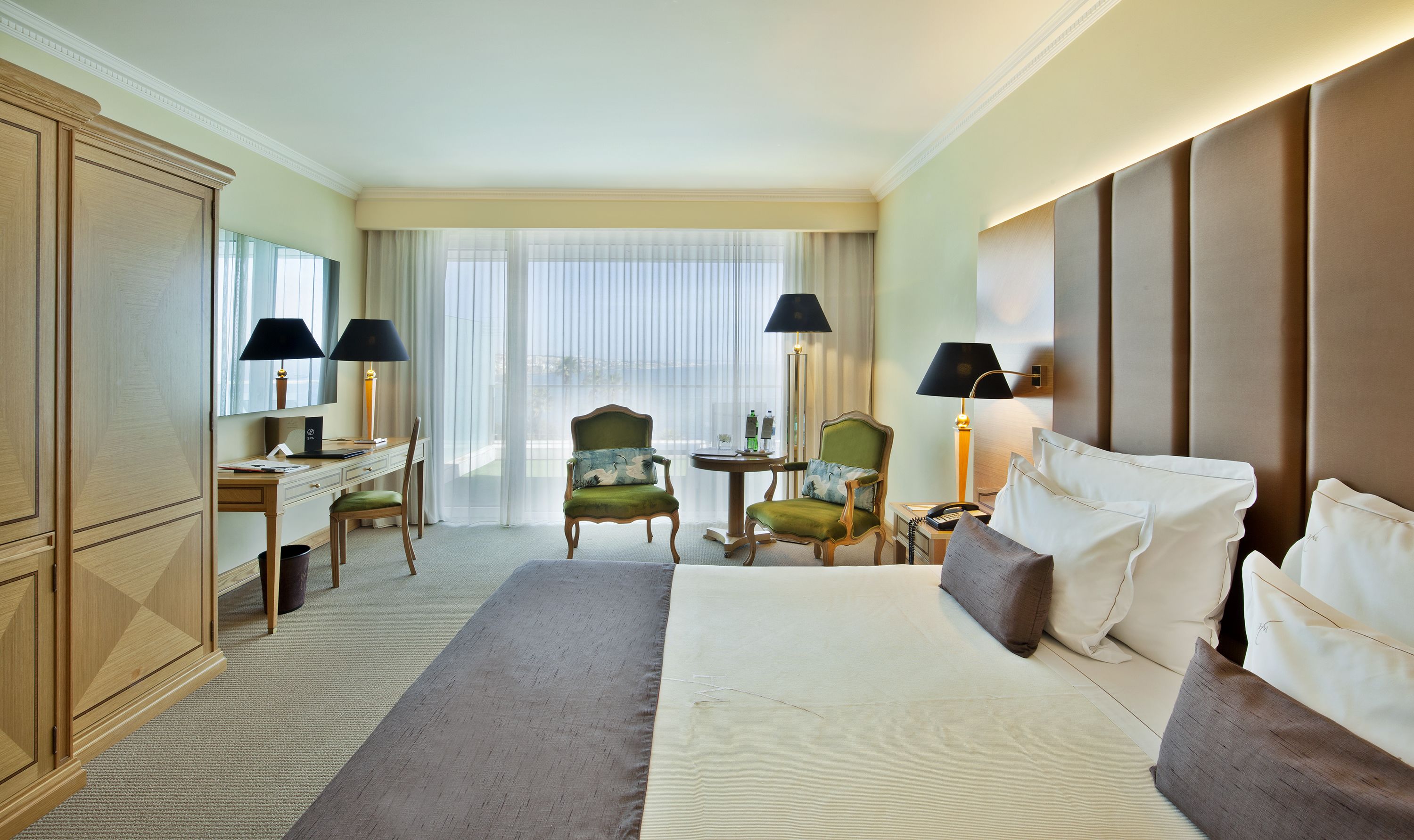 A hotel room with a large bed, two armchairs, a desk, a flat-screen TV, and a large window with sheer curtains.