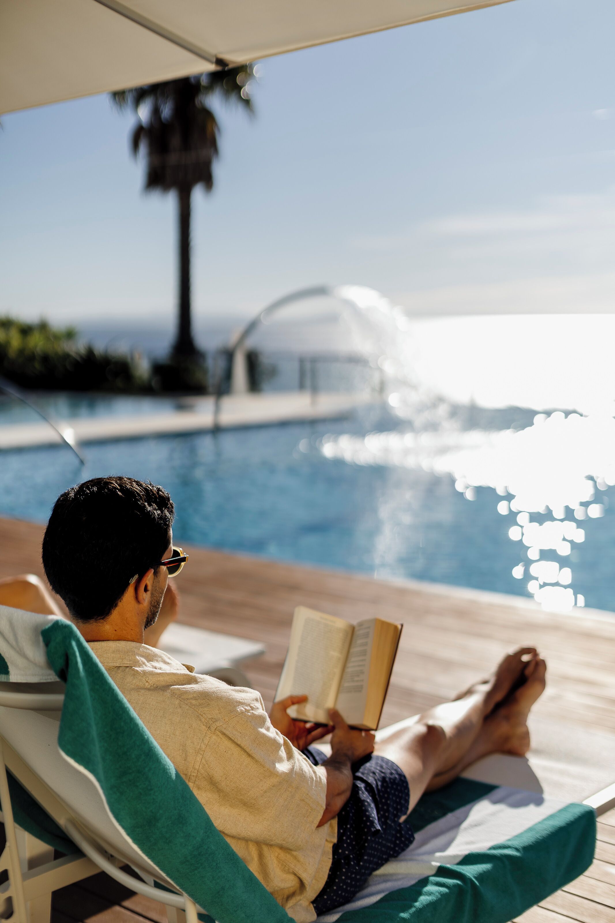 Eine Person entspannt am Pool, liest ein Buch und genießt den Blick auf das Wasser. Im Hintergrund steht eine Palmenbaum.