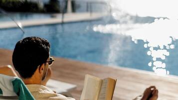 Una persona relajándose junto a una piscina con un libro, vista despejada del agua y una palmera al fondo.