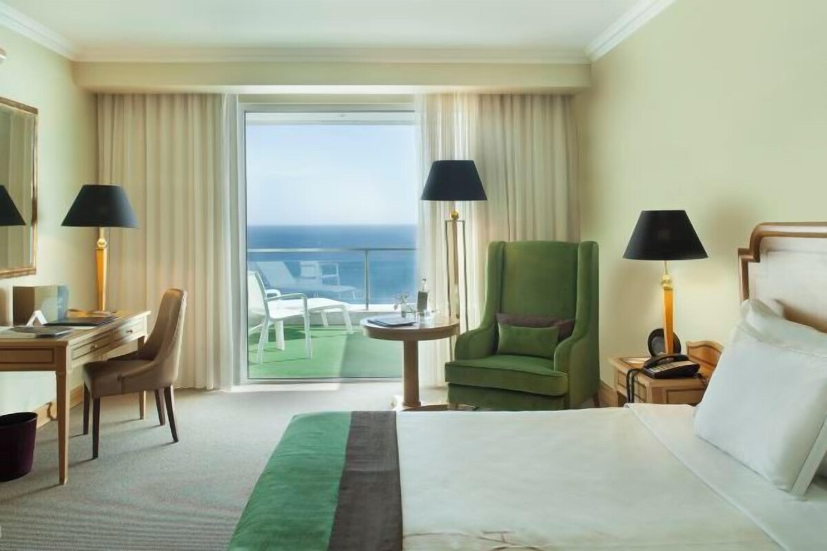 Habitación de hotel con cama, escritorio, silla, lámparas y vista al océano.