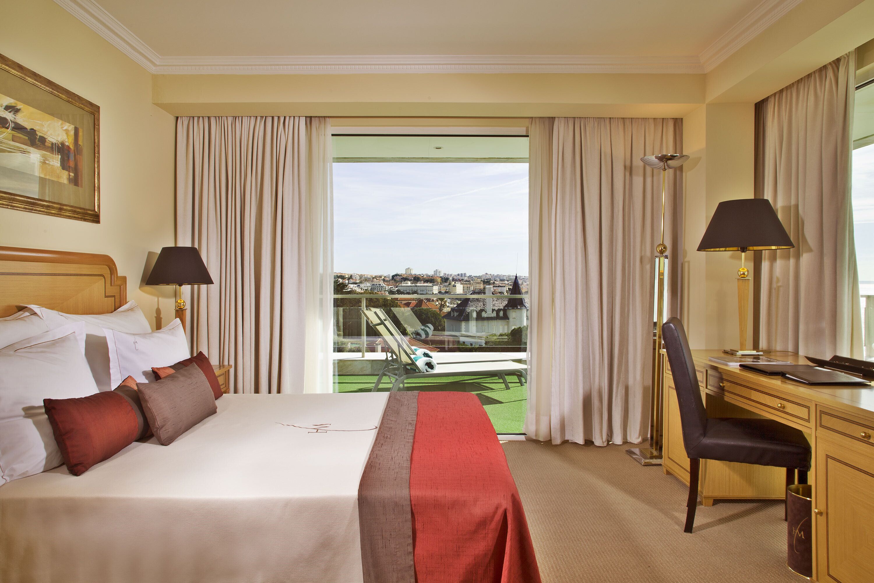 A hotel room with a bed, desk, and chair, a city view through the window, and a balcony with a railing.