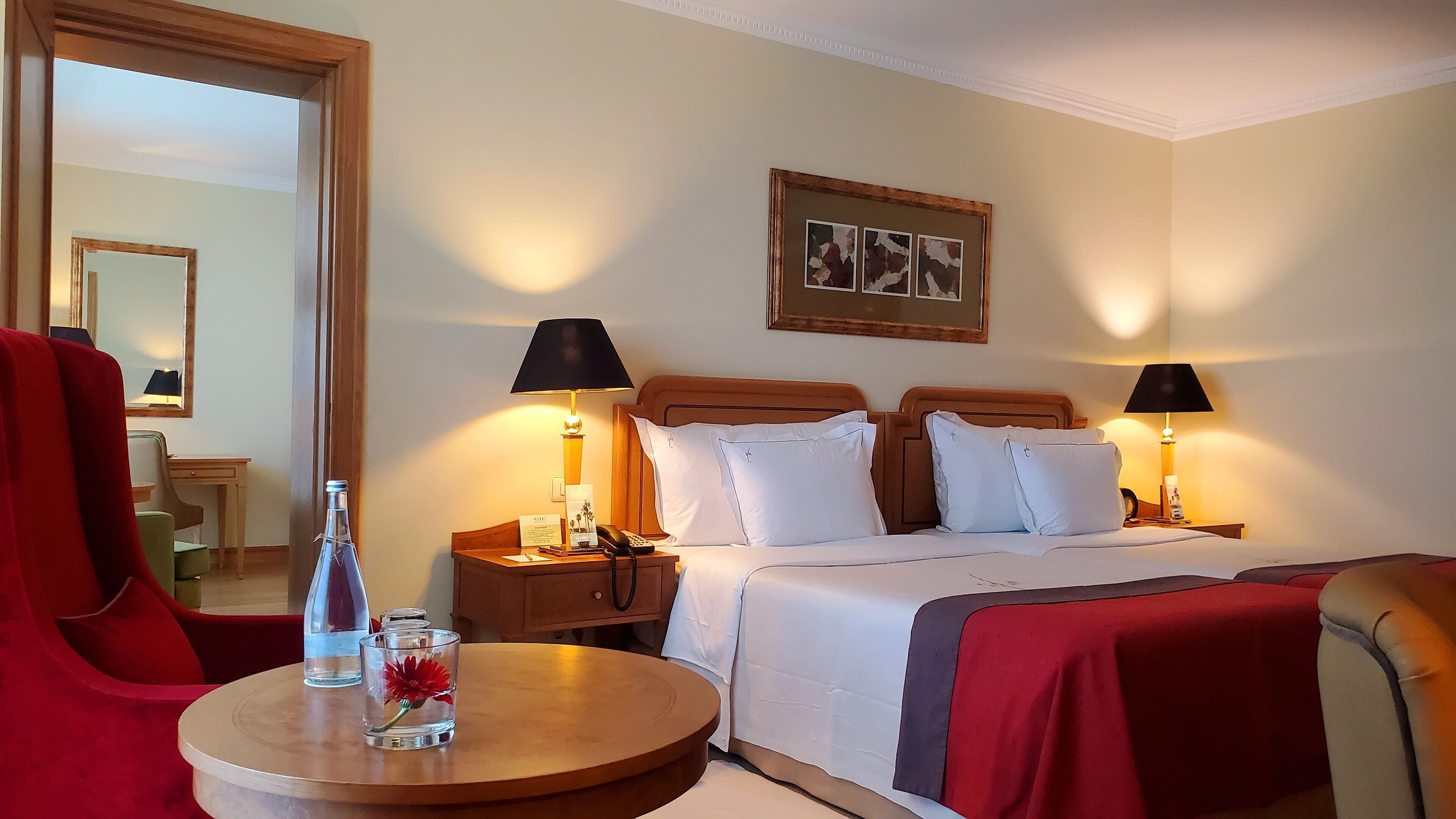 Een hotelkamer met een bed, twee nachtkastjes met lampen, een bureau, een stoel en een ronde tafel met een fles en een glas.