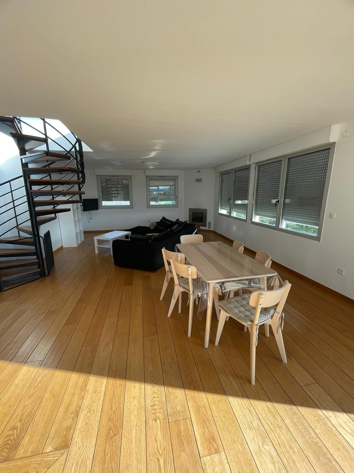 A spacious living area with a wooden floor, a dining table with chairs, a black sofa, and a spiral staircase.