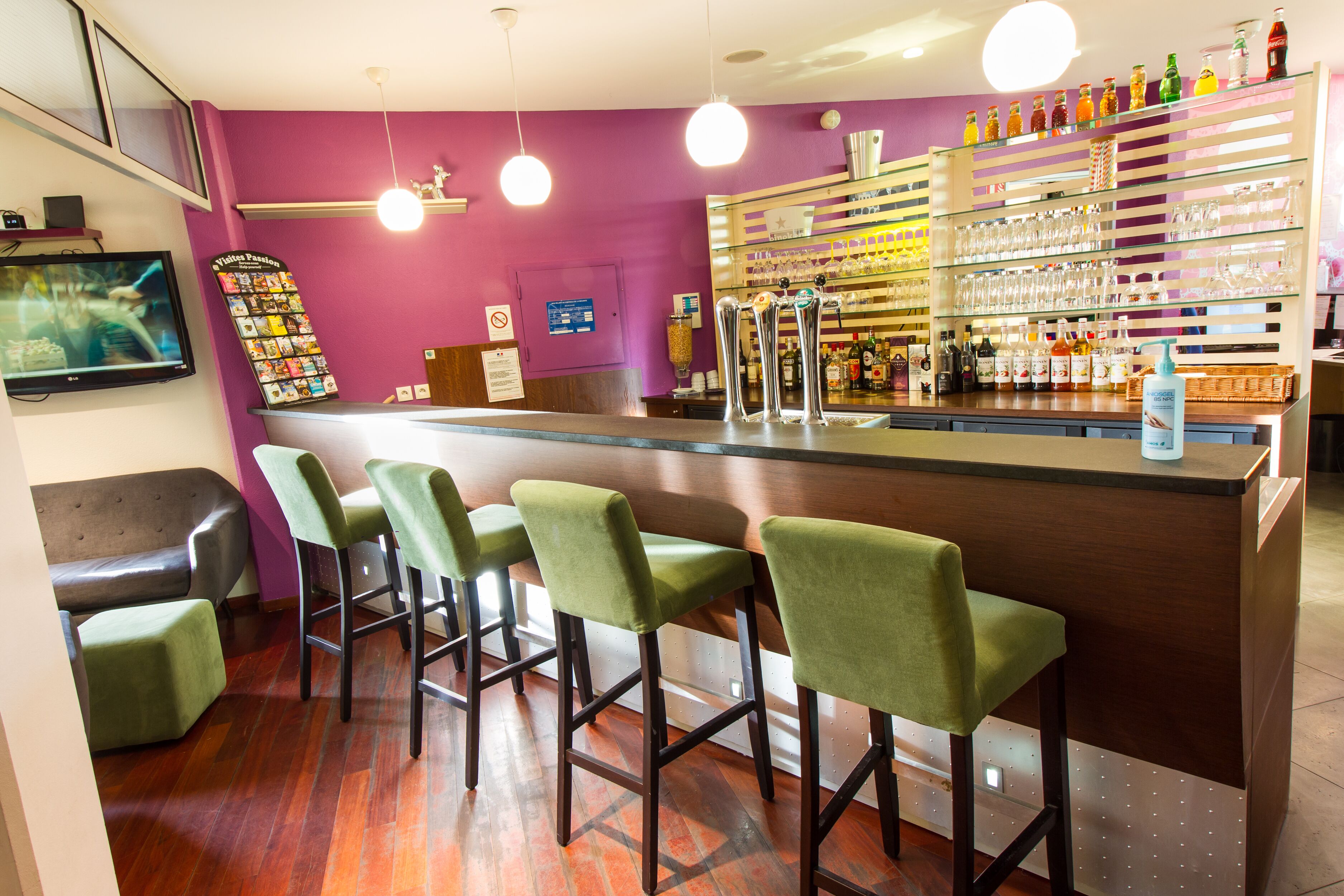 A bar area with a purple wall, green bar stools, and a variety of bottles and glasses on shelves.