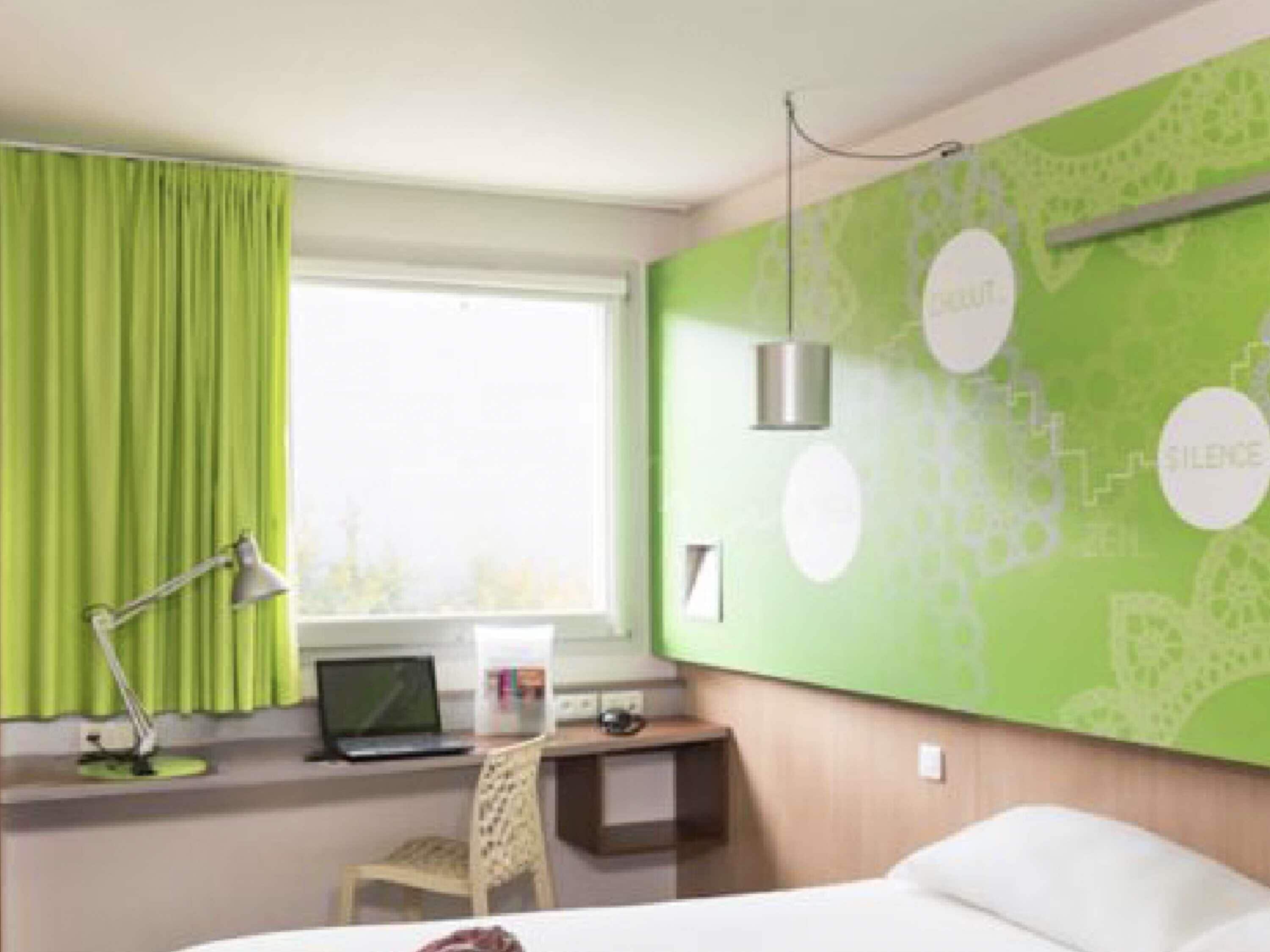 A hotel room with a bed, a desk with a laptop, a chair, a lamp, and a window with curtains.