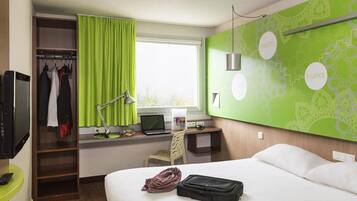 A hotel room with a bed, a desk with a laptop, a TV, a wardrobe, and a green wall with circular designs.