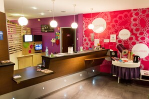 Een receptie met een houten balie, een paarse muur met de borden 'WELKOM' en 'ENVIRONMENT', een televisie en een tafel met een koffiezetapparaat en bekers.